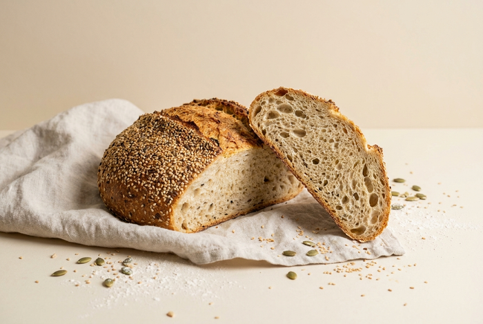 A round seeded gluten-free loaf with one thick slice cut, on linen over a beige background